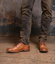 A person wearing Bed Stu Fierce men’s derby shoes with a stylish finish and dark jeans stands on a worn concrete floor against a distressed wall, showcasing the classic Goodyear leather welt.