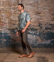 A man stands before a textured brick wall, wearing a patterned short-sleeve shirt, dark jeans, and Bed Stu's Fierce brown men’s derby shoes with a stylish finish.