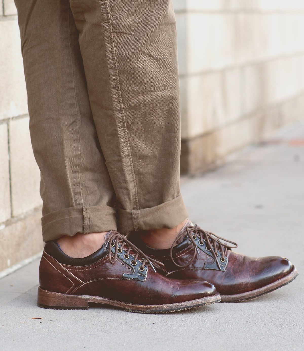 A person wearing Bed Stu's Fierce brown leather Derby shoes with a classic design and rolled-up khaki pants stands on a sidewalk, effortlessly blending modern flair.