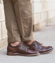 A person wearing Bed Stu's Fierce brown leather Derby shoes with a classic design and rolled-up khaki pants stands on a sidewalk, effortlessly blending modern flair.