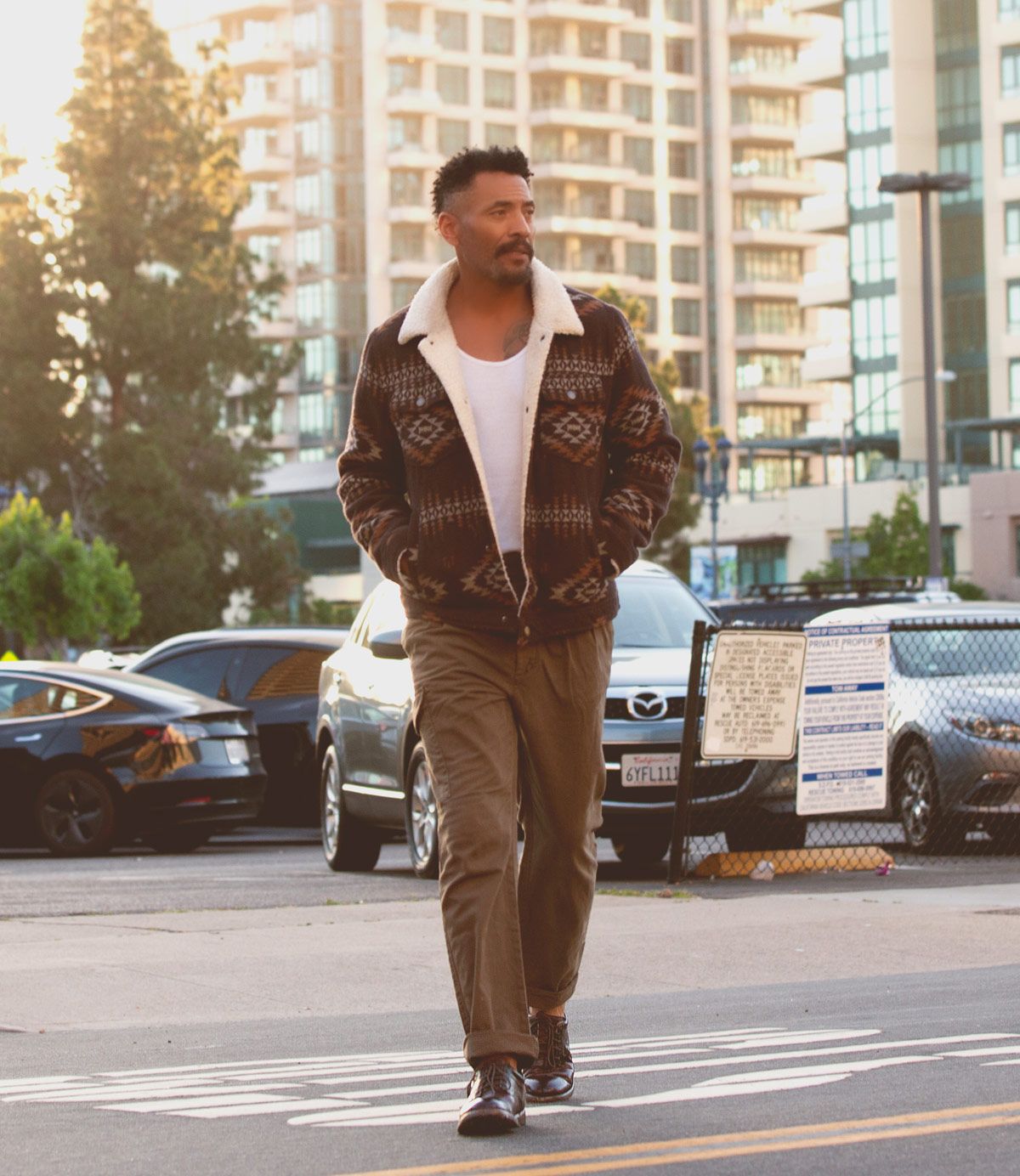 A man, exuding a modern flair, strolls through a parking lot with tall buildings as his backdrop. He's clad in a patterned jacket and white shirt, complemented by brown pants and polished men's derby shoes from the "Fierce" collection by Bed Stu.