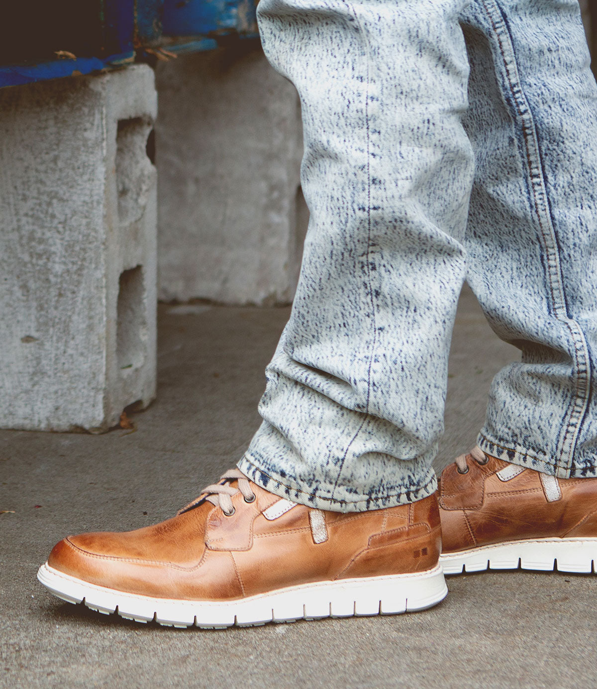 A person wearing light blue jeans and brown Bed Stu Galahad high-top leather sneakers with breathable insoles stands on a concrete surface near some cinder blocks.