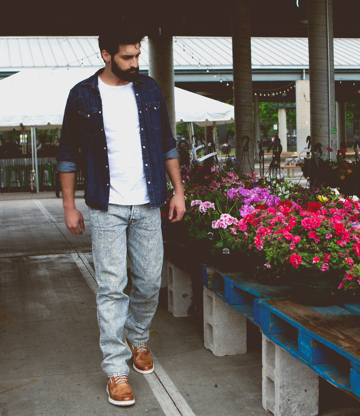 A man in a white t-shirt, denim shirt, jeans, and high-top leather sneakers with a breathable insole looks at flowers at an outdoor market wearing Galahad by Bed Stu.