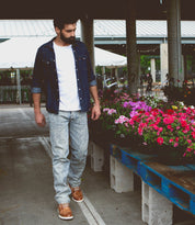 A man in a white t-shirt, denim shirt, jeans, and high-top leather sneakers with a breathable insole looks at flowers at an outdoor market wearing Galahad by Bed Stu.