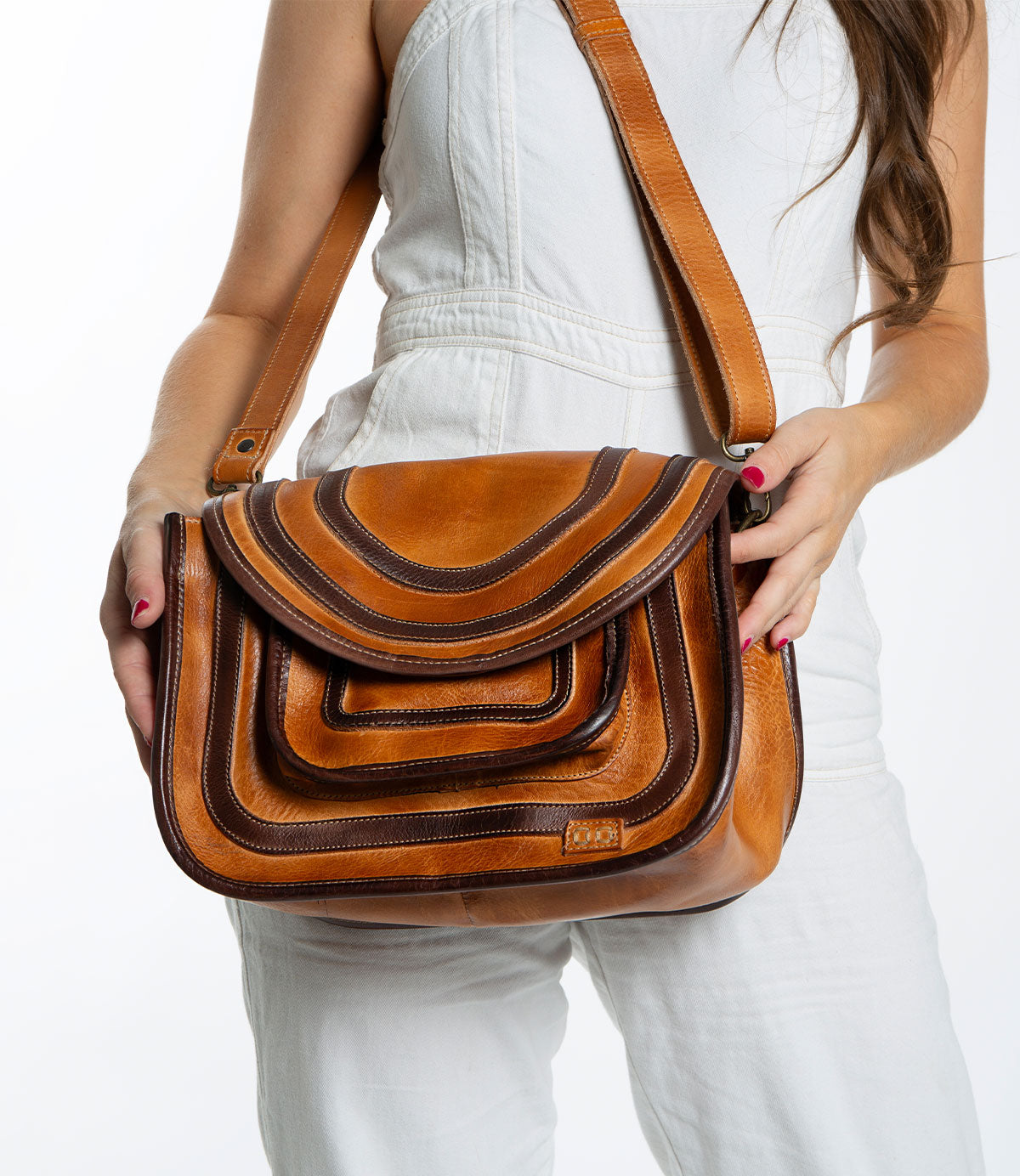 A person in a white outfit holds the Gilda bag by Bed Stu, a spacious brown leather accessory featuring a layered flap design and dual-finished leather.