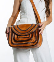A person in a white outfit holds the Gilda bag by Bed Stu, a spacious brown leather accessory featuring a layered flap design and dual-finished leather.