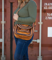 A person in a green top and jeans holds the Bed Stu Gilda, a brown striped dual-finished leather shoulder bag, in front of a shipping container.