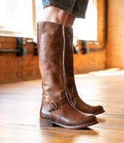 A person wearing Bed Stu Glaye knee-high brown leather boots with vintage buckle details stands indoors on a wooden floor.