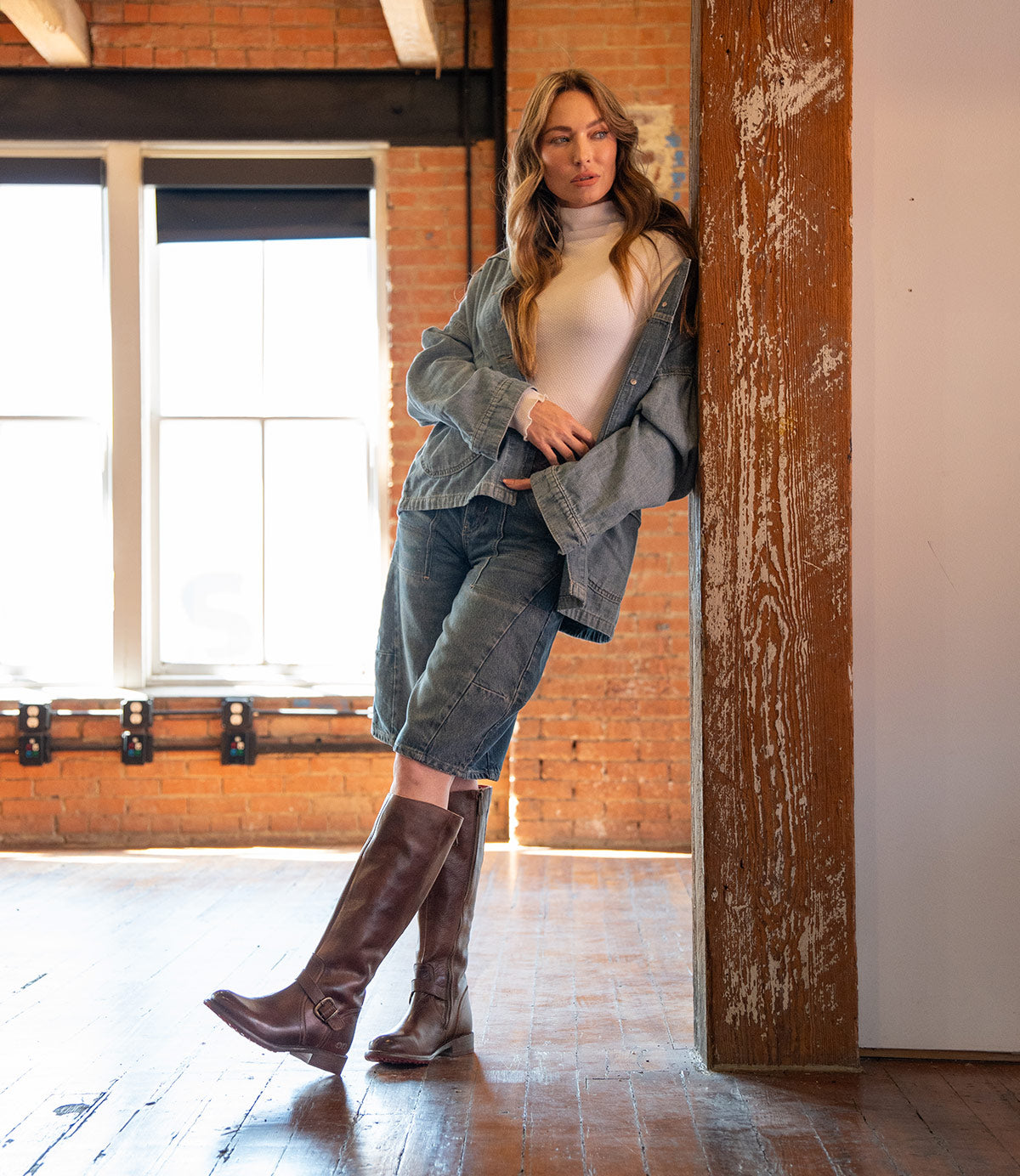 A woman wearing Bed Stu's Glaye brown equestrian boots with a vintage buckle, denim shorts, a jean jacket, and a white turtleneck leans against a wooden pillar in a sunlit room with brick walls and large windows.