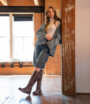 A woman wearing Bed Stu's Glaye brown equestrian boots with a vintage buckle, denim shorts, a jean jacket, and a white turtleneck leans against a wooden pillar in a sunlit room with brick walls and large windows.