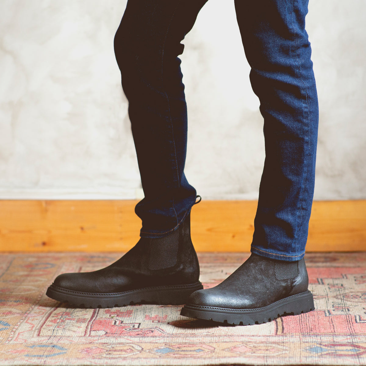 A man exuding casual elegance, standing on a rug in Bed Stu Grit Chelsea boots of enduring quality.