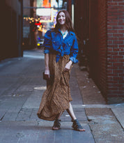 Wearing the stylish Hera gladiator sandals by Bed Stu, a woman confidently struts down a brick alleyway, her smile shining as she complements her outfit with a denim jacket and leopard print skirt.
