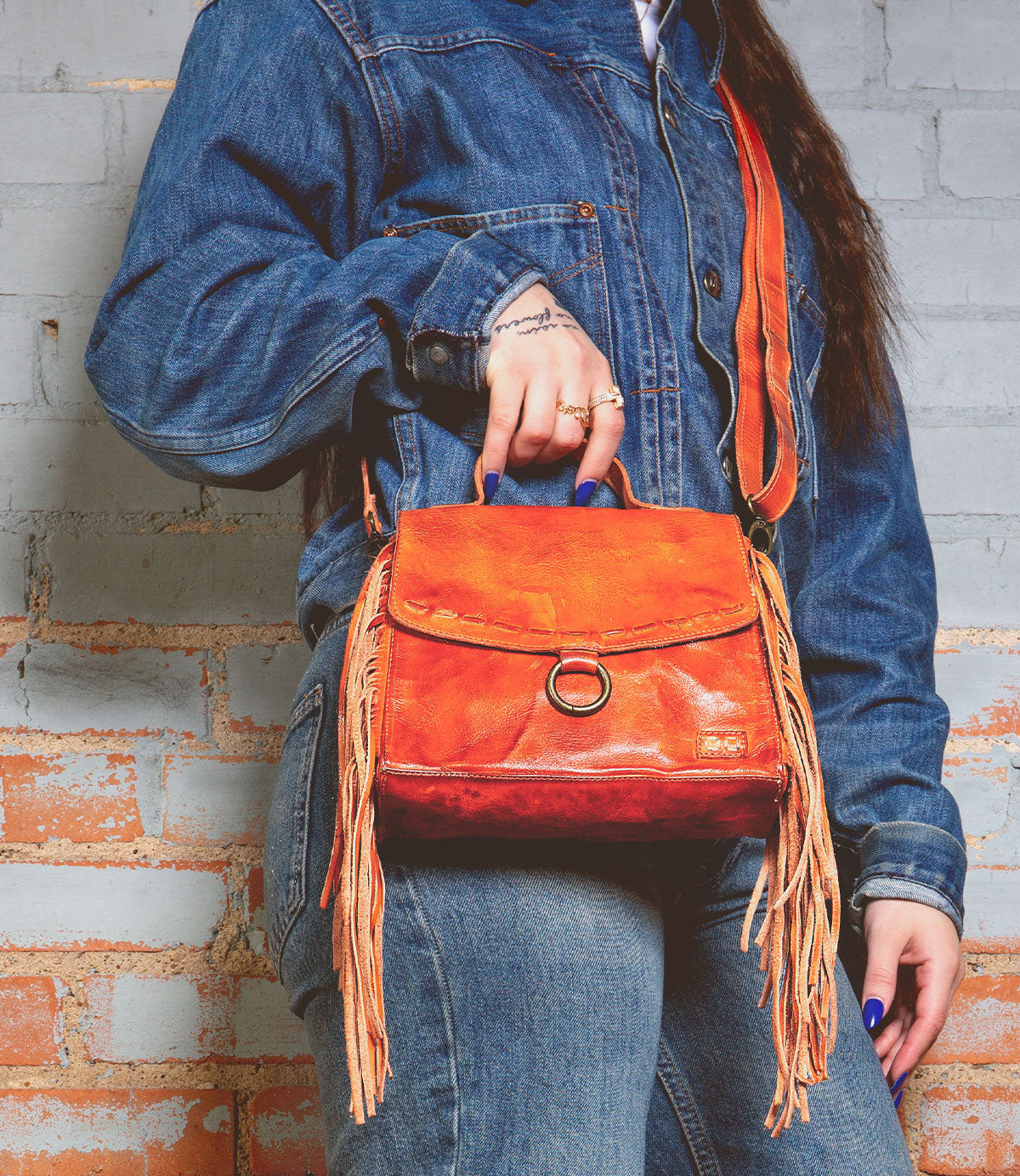 A person in a denim jacket holds a Bed Stu crossbody bag crafted from orange leather, featuring fringe accents and an adjustable strap.