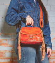A person in a denim jacket holds a Bed Stu crossbody bag crafted from orange leather, featuring fringe accents and an adjustable strap.