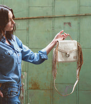 A woman holding a White Leather Crossbody Bag by Bed Stu in front of a green wall.