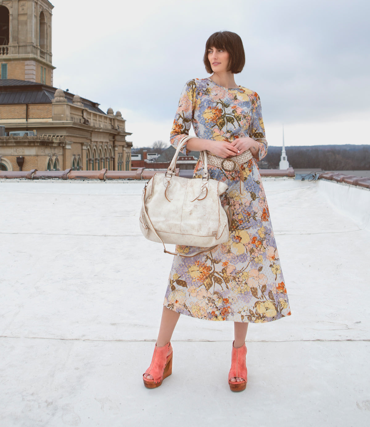 Woman in a floral dress and red shoes with ankle buckles holding a large white "Imelda" handbag on a rooftop with a cloudy sky backdrop and distant buildings. (Bed Stu)