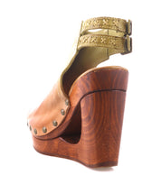 The Imelda by Bed Stu is a tan leather wedge sandal with metal studs, a thick wooden platform heel, an open toe, and two green ankle straps with decorative stitching.