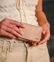 A person wearing a white textured top and light jeans holds the Bed Stu Jeor rustic leather card holder with both hands.