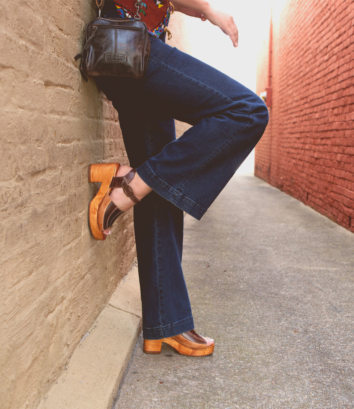A person wearing Bed Stu's Jetsetter flared jeans and brown platform shoes with patchwork finishes leaning against a brick wall.