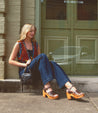 Woman sitting on a sidewalk step, smiling and looking down, wearing casual clothing with Bed Stu Jetsetter leather heels.