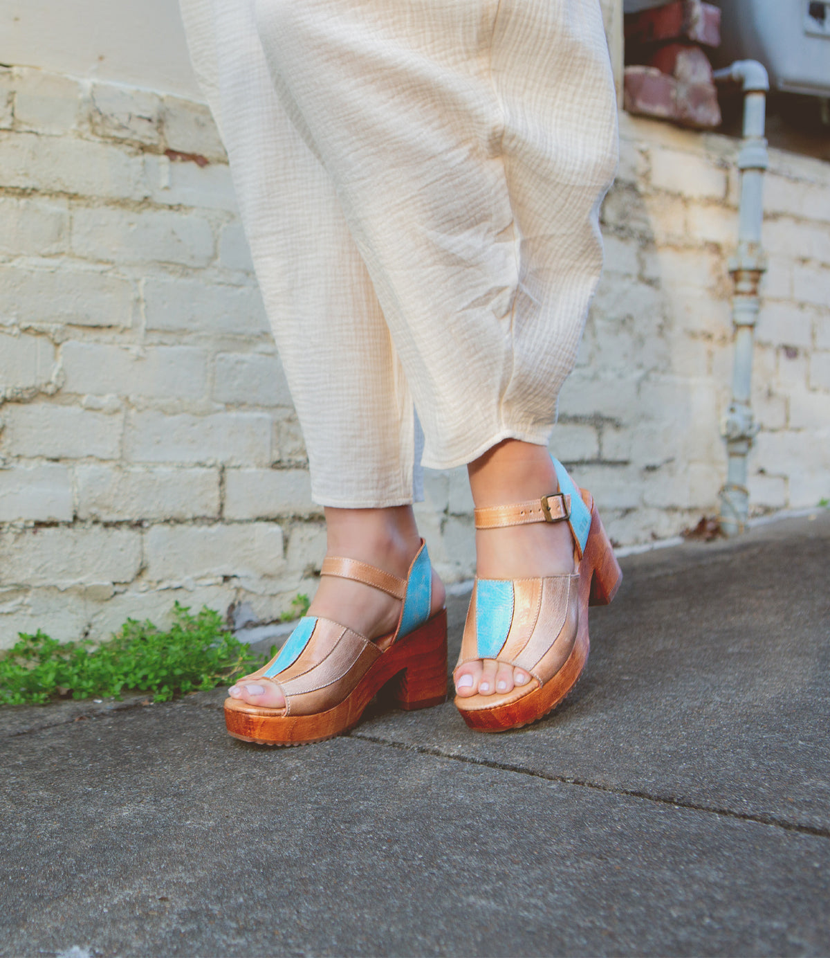 A person standing in Bed Stu's Jetsetter leather heels with patchwork finishes on a pavement next to a wall.