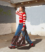 A person in a red and white striped sweater walks down the street carrying a large brown bag. They are wearing jeans and vibrant red Judgement boots by Bed Stu, featuring an 8-eye lace-up design. The cushioned insole adds comfort to their steps, while the background showcases a textured wall with patches of peeling paint.