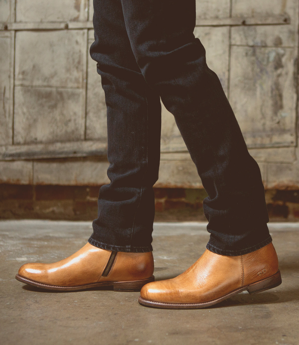 A person standing in Bed Stu Kaldi Tan Rustic Boots and dark jeans against a brick wall background.