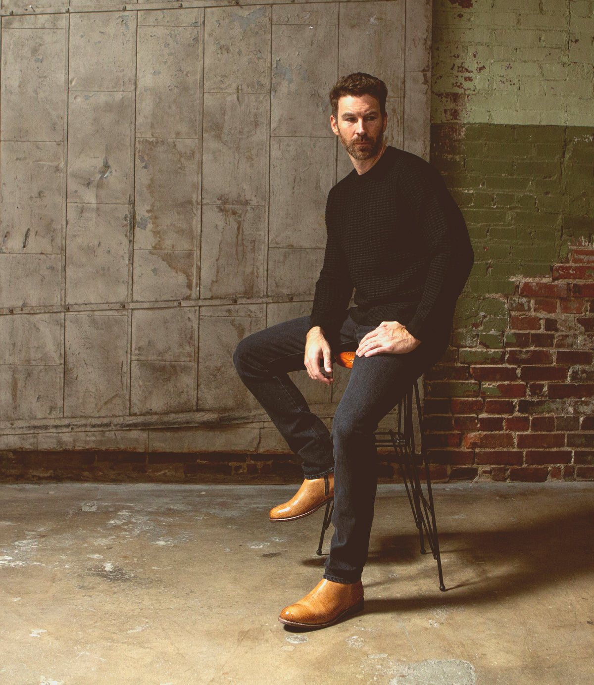 A man in a black sweater and jeans sitting on a stool in a rustic room with exposed brick walls, wearing Bed Stu leather ankle boots.