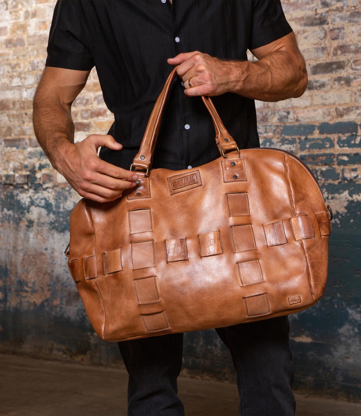 A person in a black shirt holds the Bed Stu Keg, a large tan vegetable-tanned leather duffle with a woven pattern, standing by a rustic brick wall.