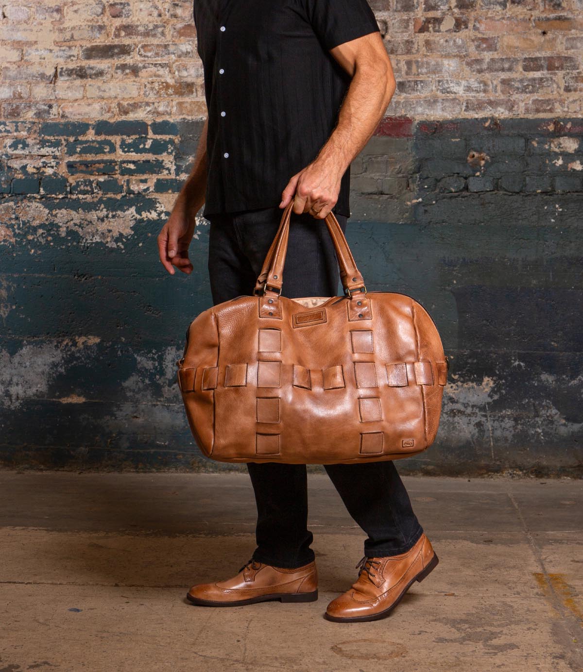 A person in a black shirt and dark pants holds the Bed Stu Keg handcrafted leather travel duffle, featuring a woven pattern, while standing against a textured brick and painted wall.