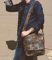 A person wearing a denim shirt and jeans holds a Bed Stu Kure with a handwoven exterior strap over their shoulder, standing against a beige concrete wall.