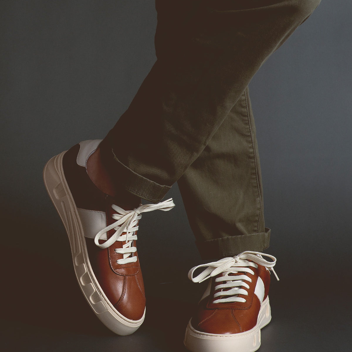 A person wearing olive green pants and Bed Stu luxury leather sneakers called Law with white laces, standing against a dark background with legs crossed.