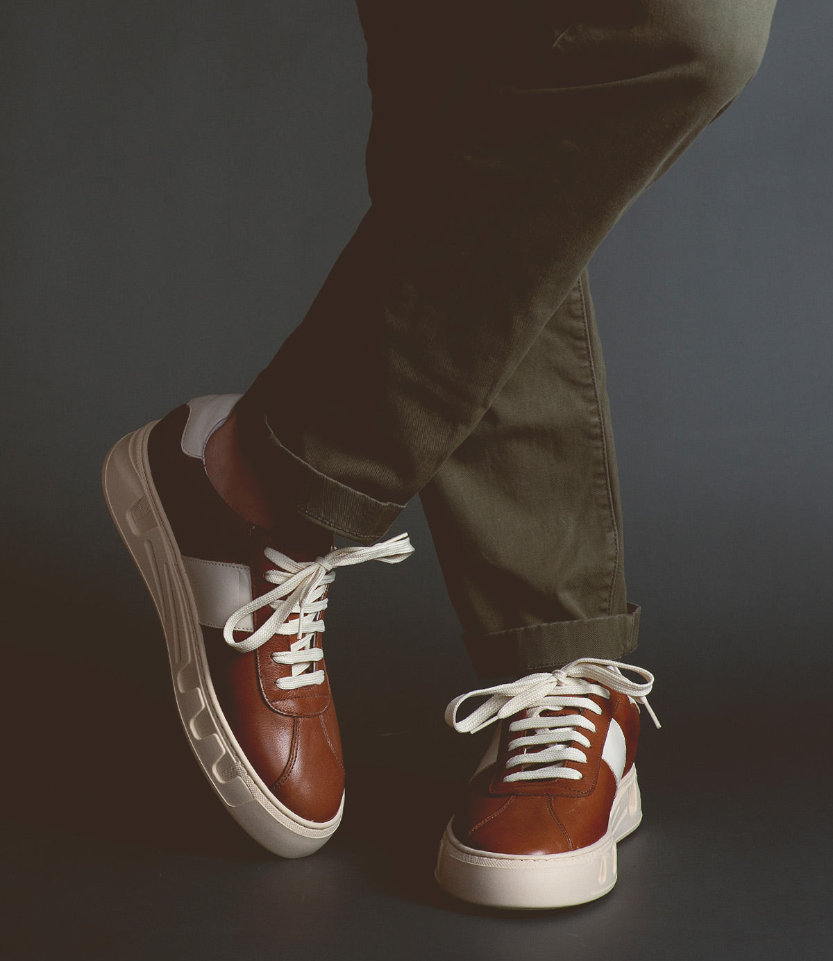 A person wearing olive green pants and Bed Stu luxury leather sneakers called Law with white laces, standing against a dark background with legs crossed.