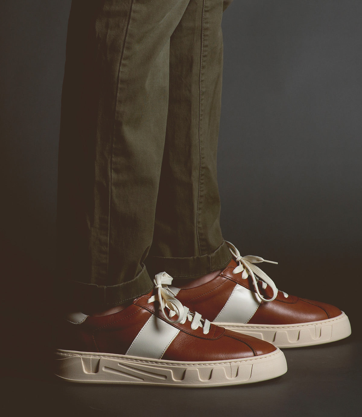 A person is wearing green pants and Bed Stu Law luxury leather sneakers with thick white soles, standing on a dark background.