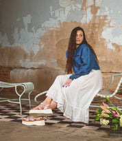 A woman in a blue top and white skirt sits on a metal chair in a rustic room, wearing Bed Stu’s Lift platform sandals—a perfect warm-weather staple—as a bouquet of flowers rests on the floor nearby.