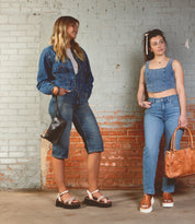Two women in denim stand against a brick wall, each carrying handbags and wearing Bed Stu Lift platform sandals with adjustable straps—a perfect warm-weather staple—as they smile at each other.