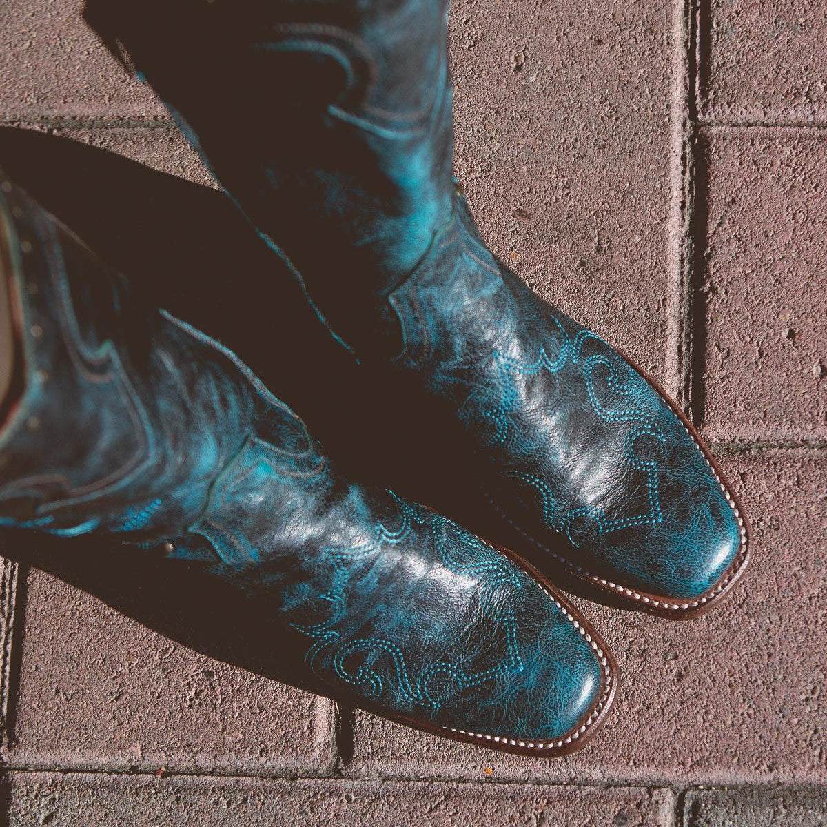 A pair of black Bed Stu Limit Western mid-calf boots with blue floral stitching, seen from above, standing on red brick pavement.