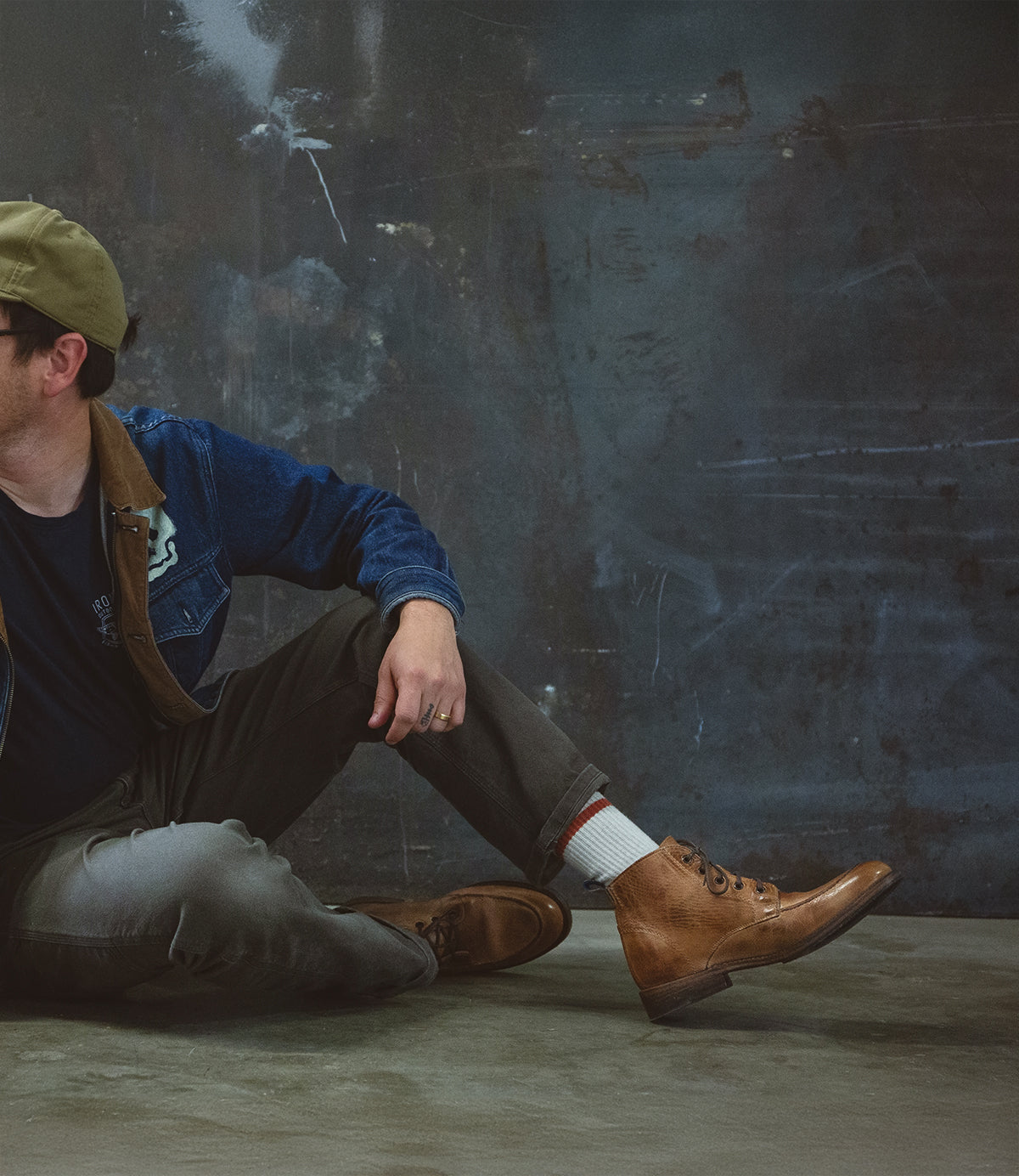 A person is sitting on the floor against a textured gray wall, wearing a green cap, denim jacket, dark pants, and a pair of sturdy Bed Stu's Lincoln II work boots with cushioned insoles.