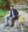 A man sitting on a rocky outcrop wearing a denim jacket and jeans, complemented by Bed Stu men's leather boots, with a contemplative expression.