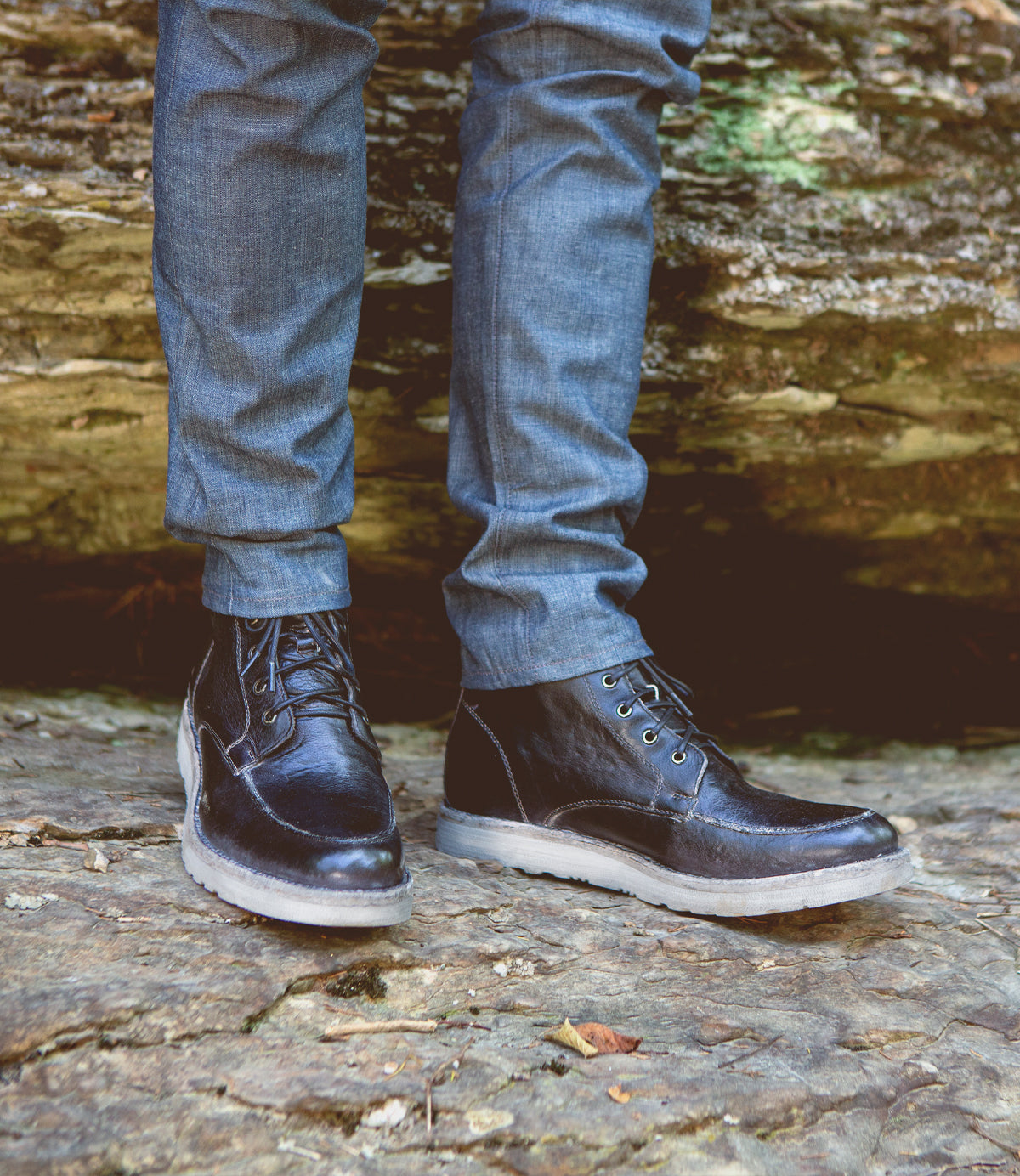 A person wearing dark jeans and a Bed Stu Lincoln Black Lux Boot standing on a rocky surface.