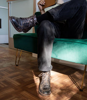 A person sitting on a green sofa with their legs crossed is holding a notebook. They are wearing jeans and the Lincoln II black lace-up boots by Bed Stu. Sunlight shines on the floor, emphasizing the comfort provided by the breathable insoles.