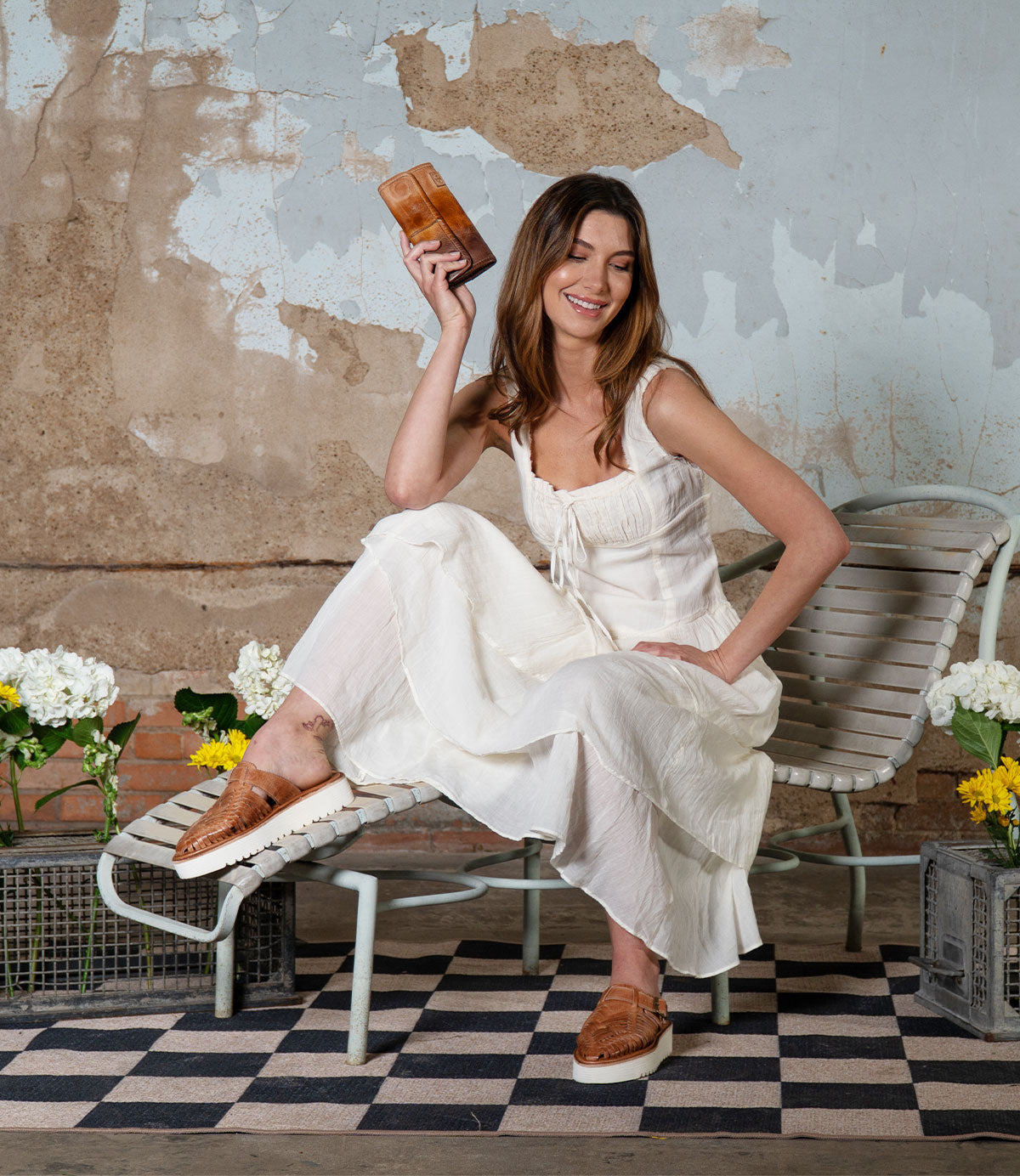 Dressed elegantly in a white dress, a woman sits gracefully on a chair, holding a small brown clutch. Her feet sport stylish Bed Stu Linette platform slides with an XL EXTRALIGHT® outsole. Surrounded by flowers against a textured wall, her look is both elegant and effortlessly chic.