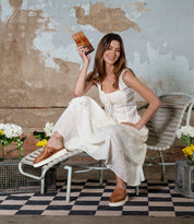 Dressed elegantly in a white dress, a woman sits gracefully on a chair, holding a small brown clutch. Her feet sport stylish Bed Stu Linette platform slides with an XL EXTRALIGHT® outsole. Surrounded by flowers against a textured wall, her look is both elegant and effortlessly chic.