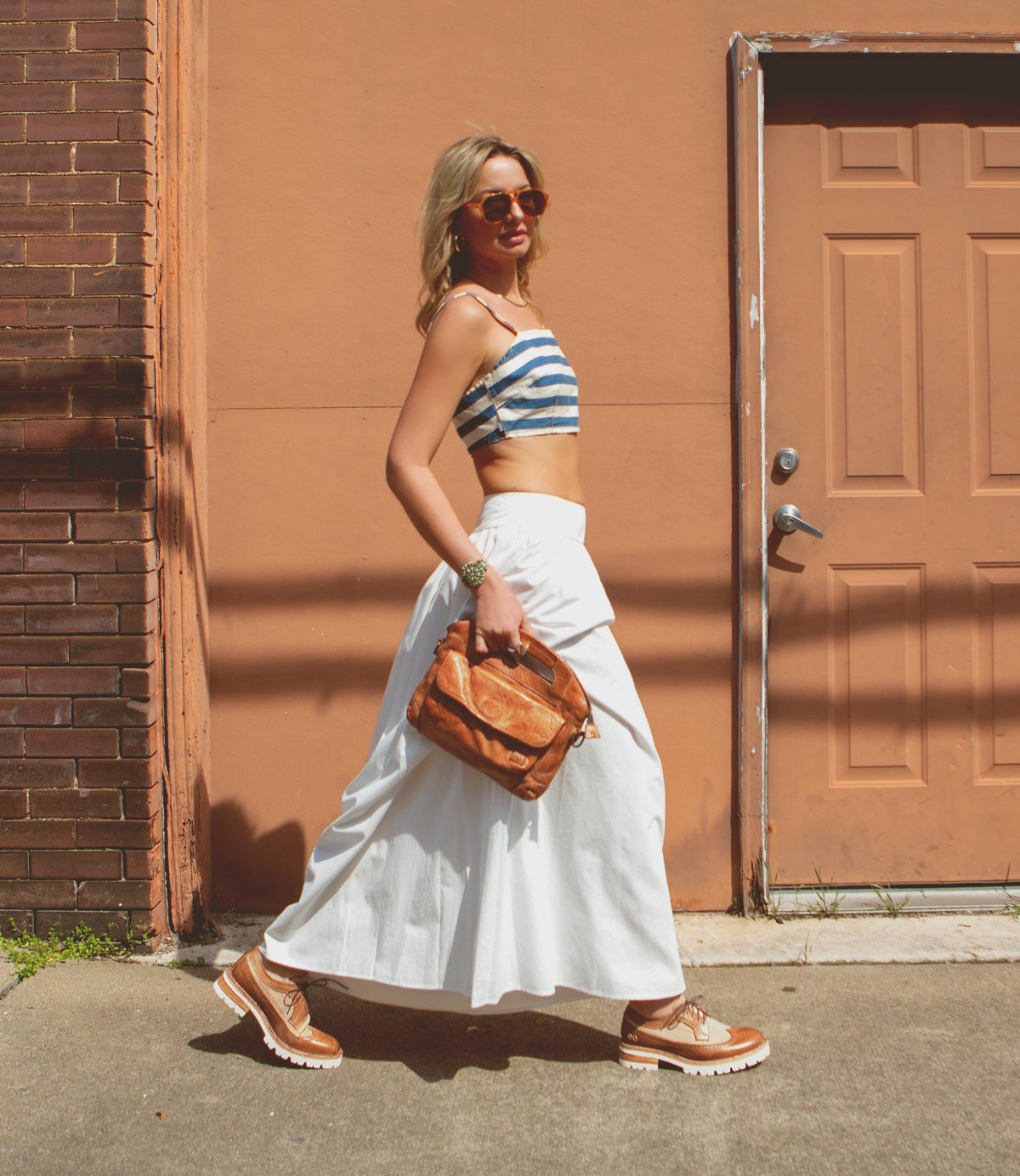 Woman in sunglasses, striped crop top, and white pants walking past an orange door with a Bed Stu Lita K III sustainable fashion brown bag in hand.