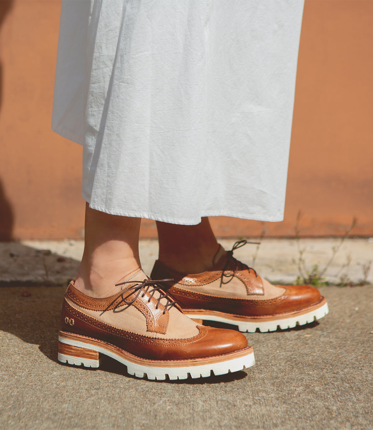 A person standing in Bed Stu Lita K III platform shoes, embodying 90’s fashion, paired with a white skirt against an orange wall.