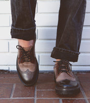 Person wearing black jeans, brown leather wingtip shoes from Bed Stu, standing on a tiled surface in front of a white brick wall.