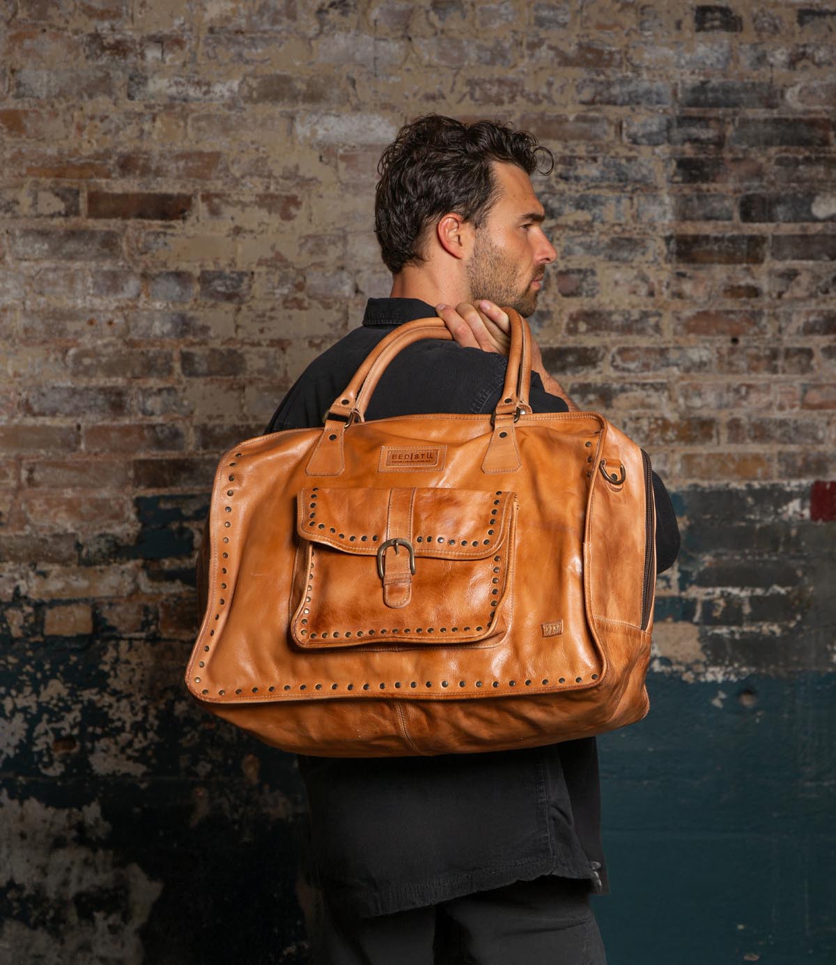 A man in a dark shirt stands against a brick wall, carrying the Bed Stu Loaf duffle bag—featuring a front pocket and stitched detailing—over his shoulder, making it a stylish carry-on ideal for weekend trips.