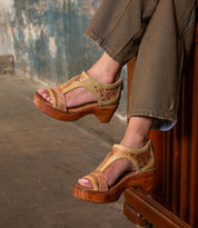 A person wearing Bed Stu's Locket vintage-inspired wooden heel sandals with cutouts and tan pants sits with crossed legs against a rustic, textured wall.