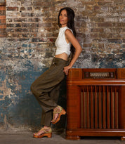 A woman in a white sleeveless top and olive pants leans against a vintage wooden radio, showcasing the Bed Stu Locket sandal with laser-cut floral details, in front of a weathered brick wall.