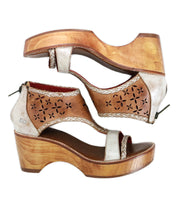 The Bed Stu Locket sandals feature vintage-inspired wooden heels, metallic straps, and laser-cut floral detailing on the sides, shown against a white background.